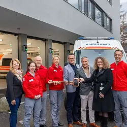 Gruppe von Menschen steht vor einem Krankenhausgebäude neben einem Krankenwagen. Sie halten eine symbolische Schlüsselübergabe ab. Schnee auf den Bergen im Hintergrund.