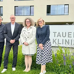 Vier Personen stehen vor einem Gebäude mit der Aufschrift "Tauern Klinikum". Sie sind geschäftlich gekleidet und lächeln in die Kamera.