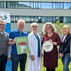 Eine Gruppe von sechs Personen steht vor einem Klinikgebäude. Sie halten Schilder und Auszeichnungen, während sie in die Kamera lächeln. Es ist sonnig.