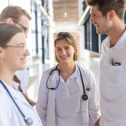 Eine Gruppe von fünf medizinischen Fachkräften in weißen Kitteln unterhält sich freundlich in einem modernen Klinikflur. Sie tragen Stethoskope.