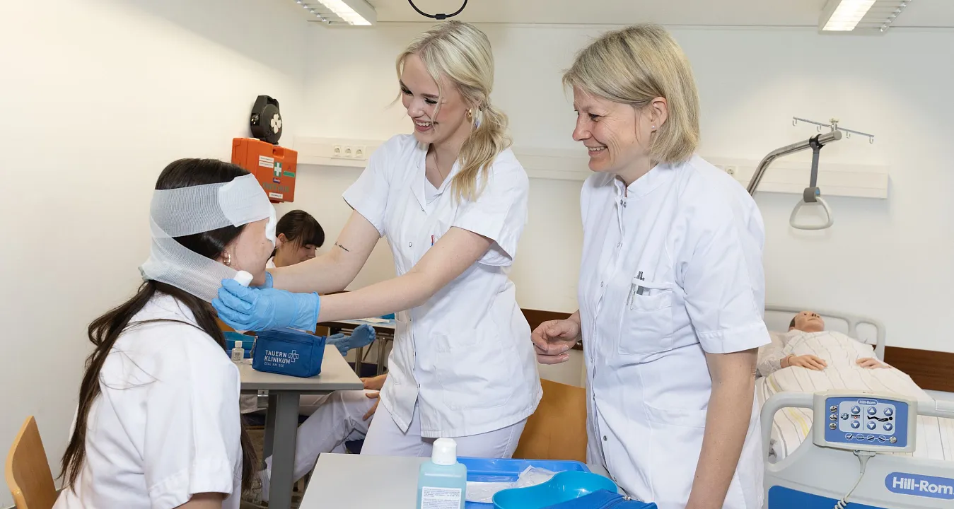 Zwei Krankenschwestern versorgen eine sitzende Patientin mit einem Kopfverband in einem Krankenhauszimmer. Im Hintergrund steht ein Bett mit einem Patienten.