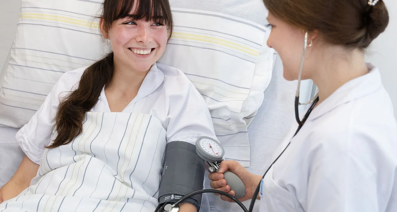 A nurse checking a patient's blood pressure. The patient is in bed, smiling, with a blood pressure cuff on her arm. Both seem relaxed and happy.